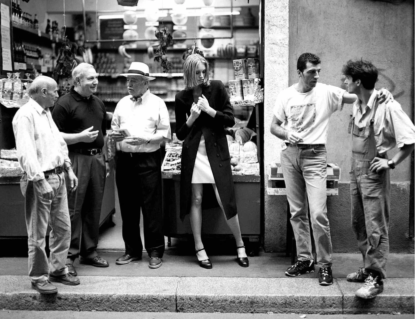 Black and white street photography of a group of people standing outside a shop with a woman in a long black coat posing.