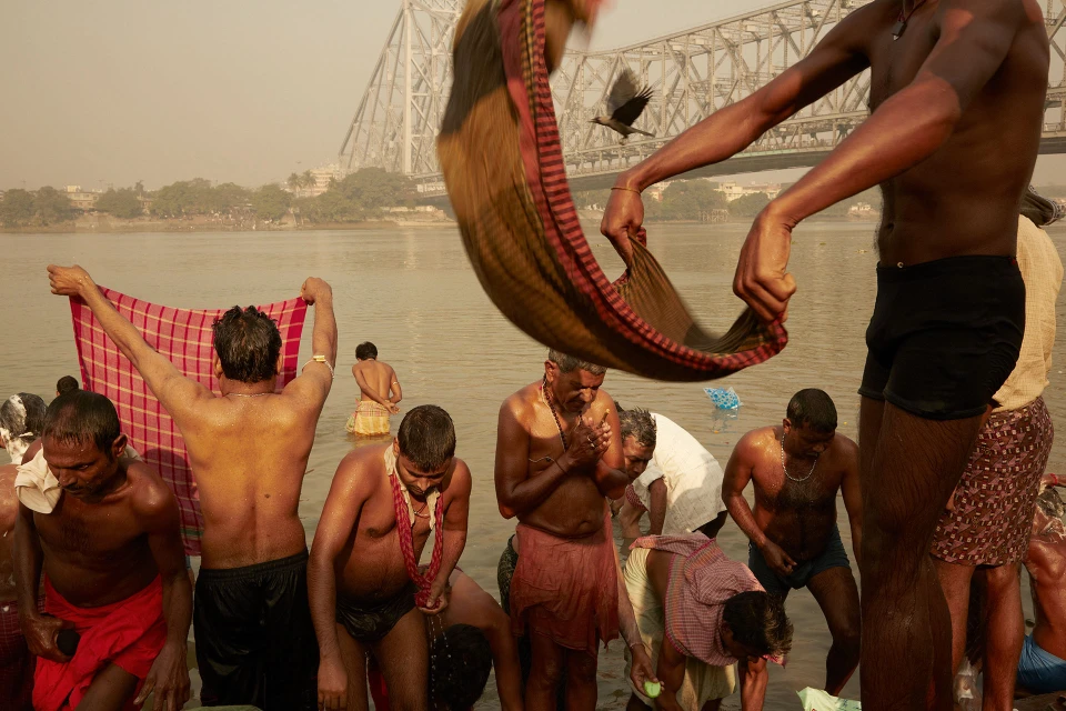 People bathe in a river at dawn in India, with a bridge in the background. Some wear colorful cloths. One person holds a towel aloft.