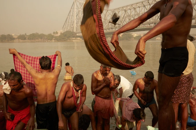 People bathe in a river at dawn in India, with a bridge in the background. Some wear colorful cloths. One person holds a towel aloft.