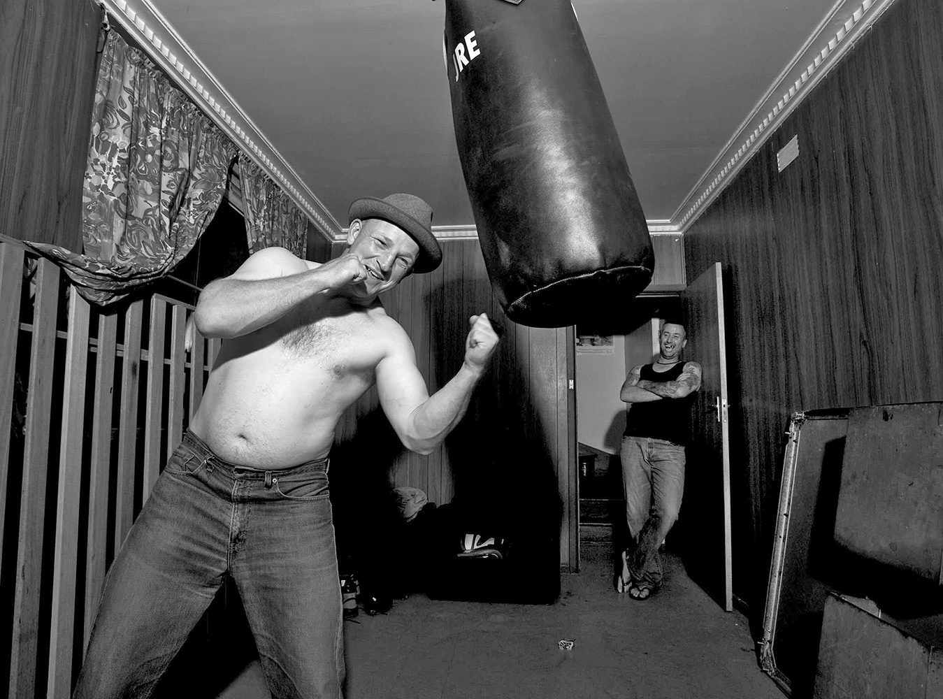 Monochrome shot of a man boxing with a punching bag indoors, with a second man observing in the background, creating a candid, gritty scene.