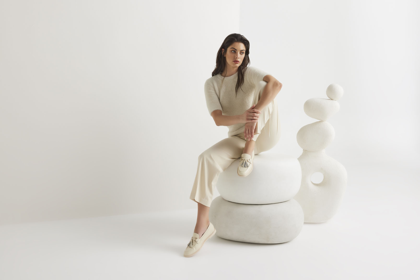 Elegant woman in cream knitwear sitting on stone-like sculpture in a bright white studio, exuding minimalistic fashion.