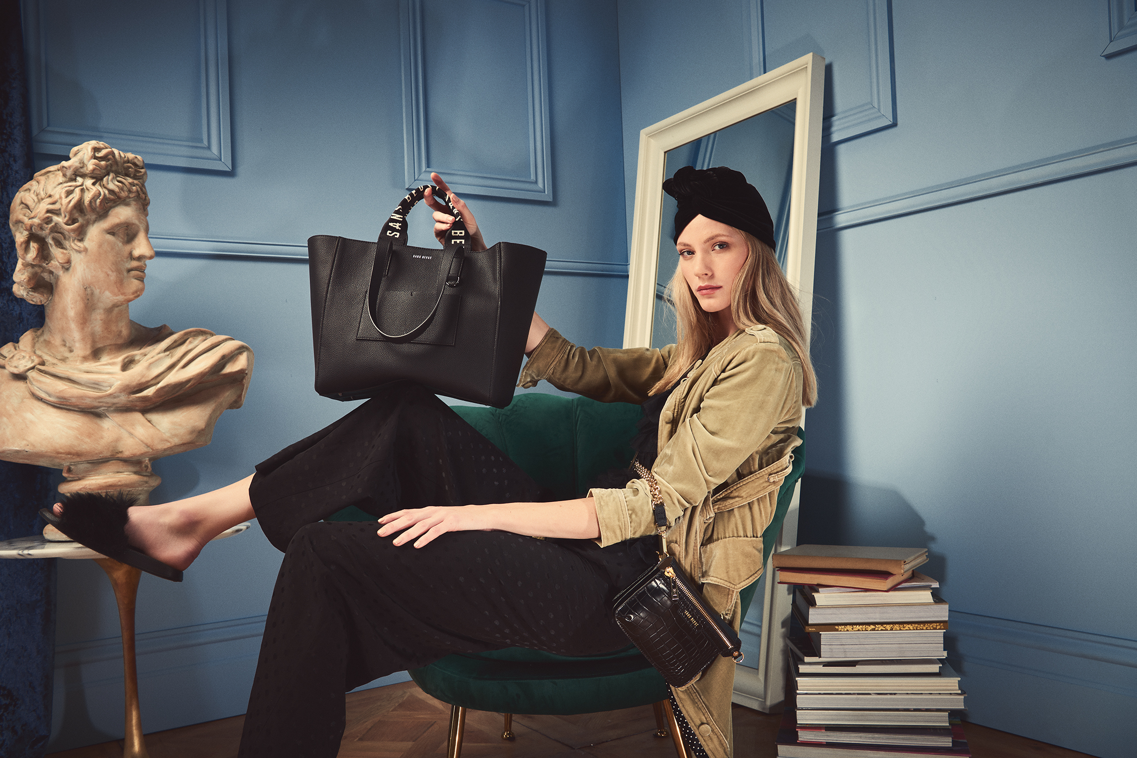A woman in a turban sits in a green chair holding a black leather purse with a sculpture and books in the background.
