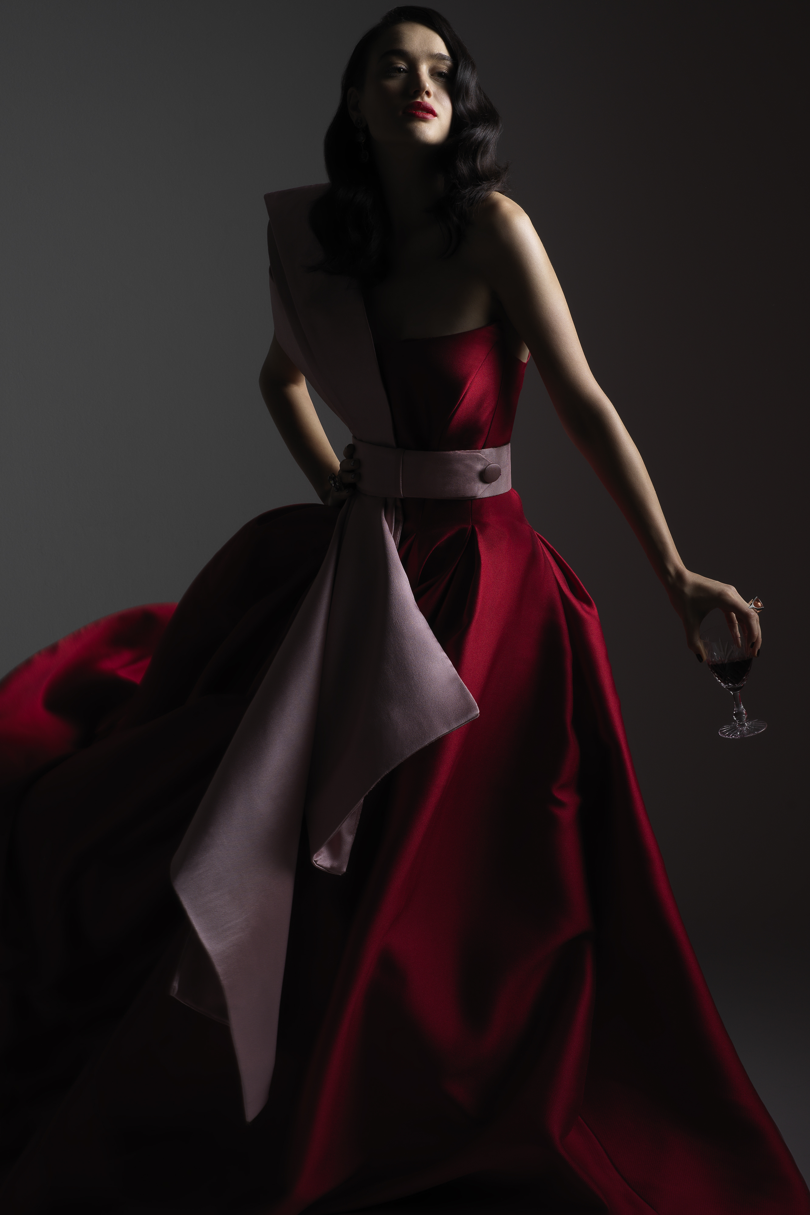 Elegant woman in red gown holds a glass of red wine, posing against a dark backdrop with soft lighting and luxurious fabric details.