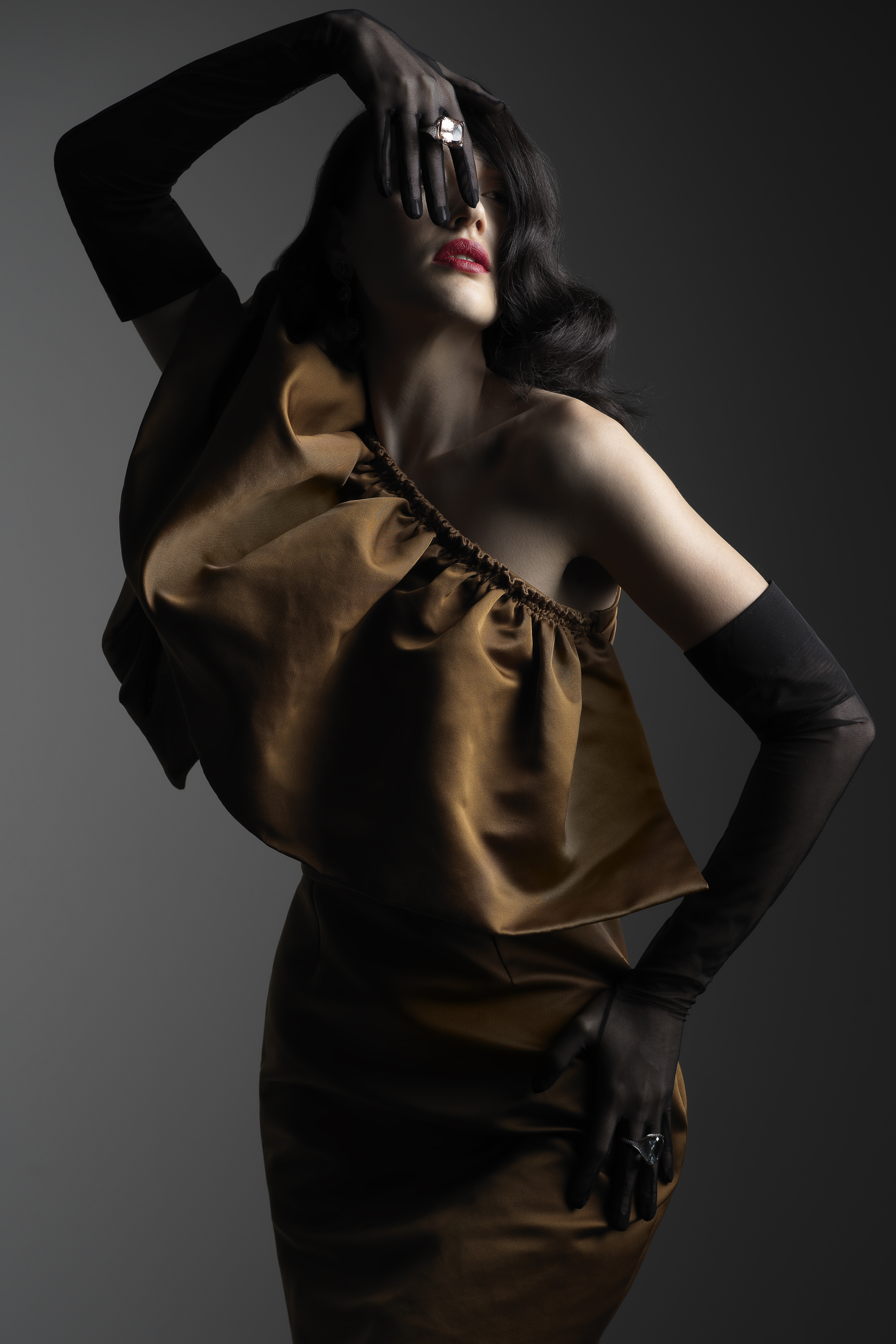 Brunette woman in gold dress and black gloves poses in a studio with hand near face, ring visible, wearing red lipstick, on gray background.