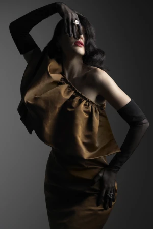 Woman in bronze satin one-shoulder ruffled top with black sheer gloves posing dramatically against a dark gradient background.