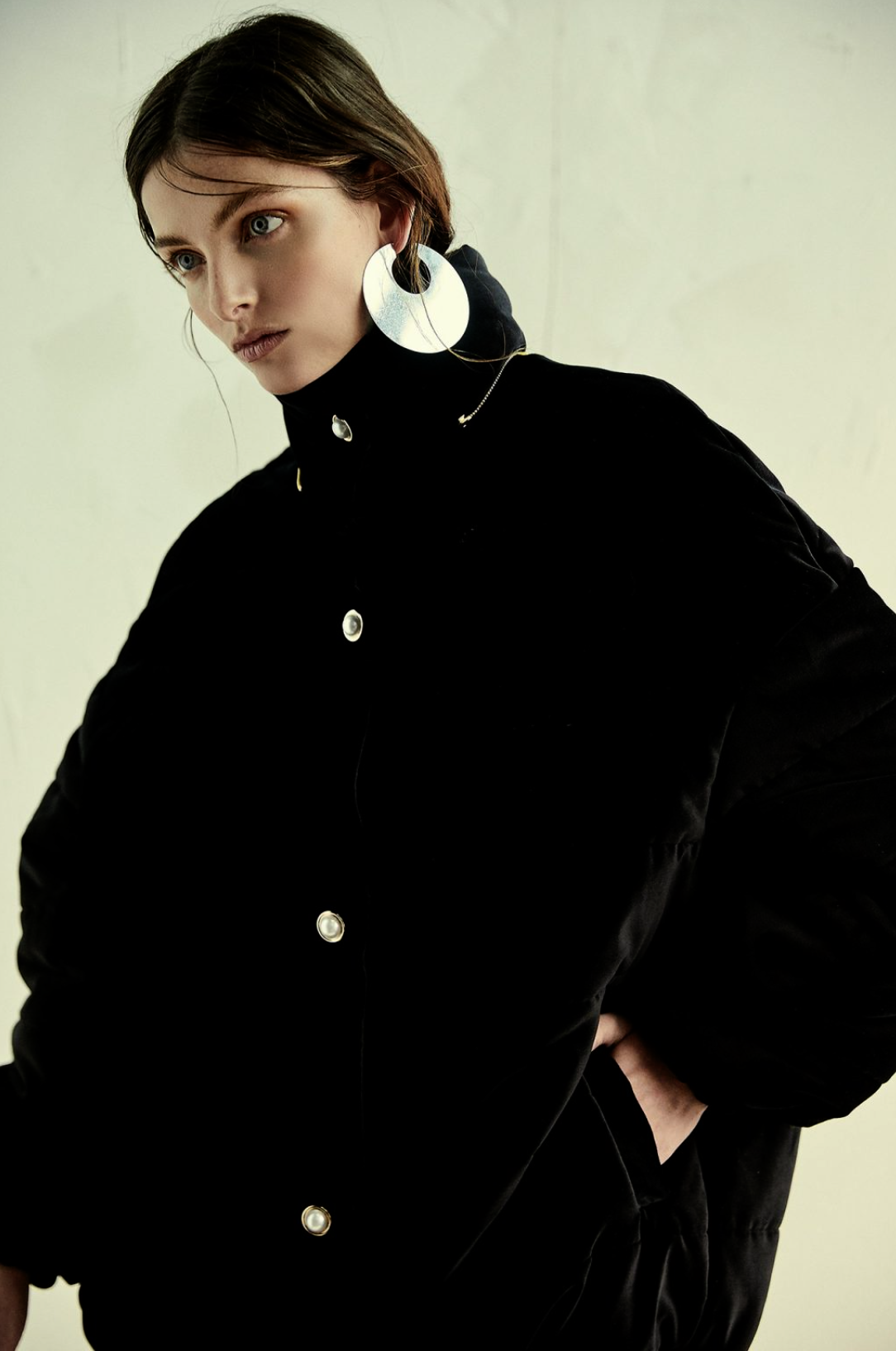 Fashion portrait of a woman with silver earrings wearing a black velvet coat against an off-white background in soft, diffused lighting.