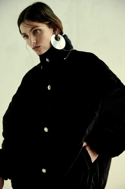Fashion portrait of a woman with silver earrings wearing a black velvet coat against an off-white background in soft, diffused lighting.
