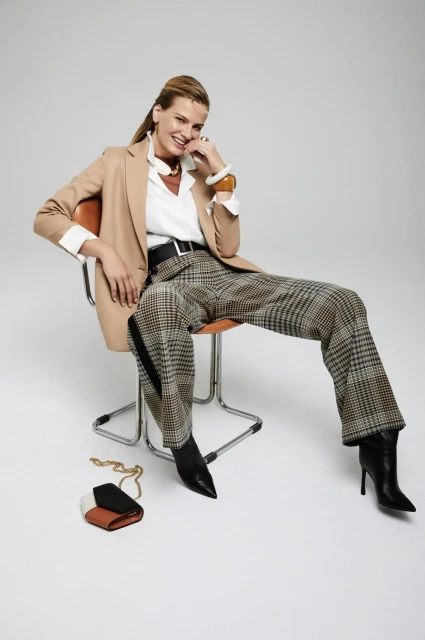 Woman in beige blazer and plaid pants sits in chair, accessorized with jewelry and bag in a light-toned studio. 