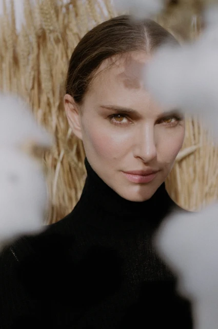 Close-up portrait of a woman with fair skin and brown hair, wearing a black turtleneck sweater, posed in front of a wheat field.