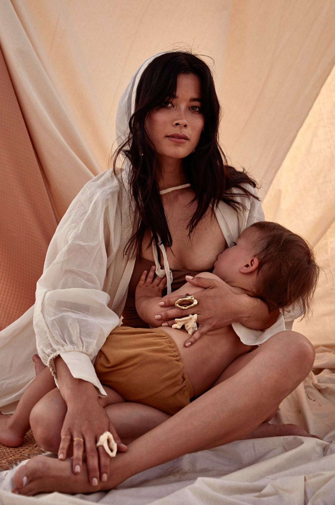 Brunette woman breastfeeding infant in cream-colored clothing and background. Soft lighting, shallow depth of field. Mother and child portrait.