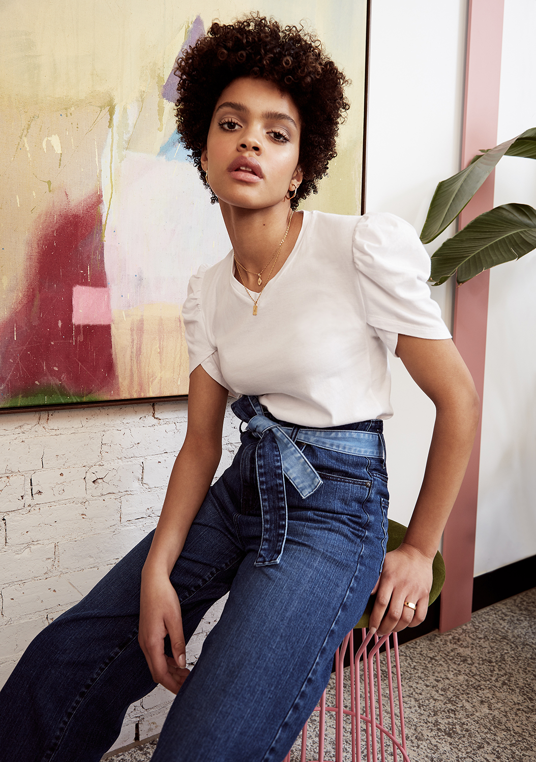 A brunette woman with an afro is sitting indoors wearing a white t-shirt, dark denim pants with a sash, and gold jewelry.