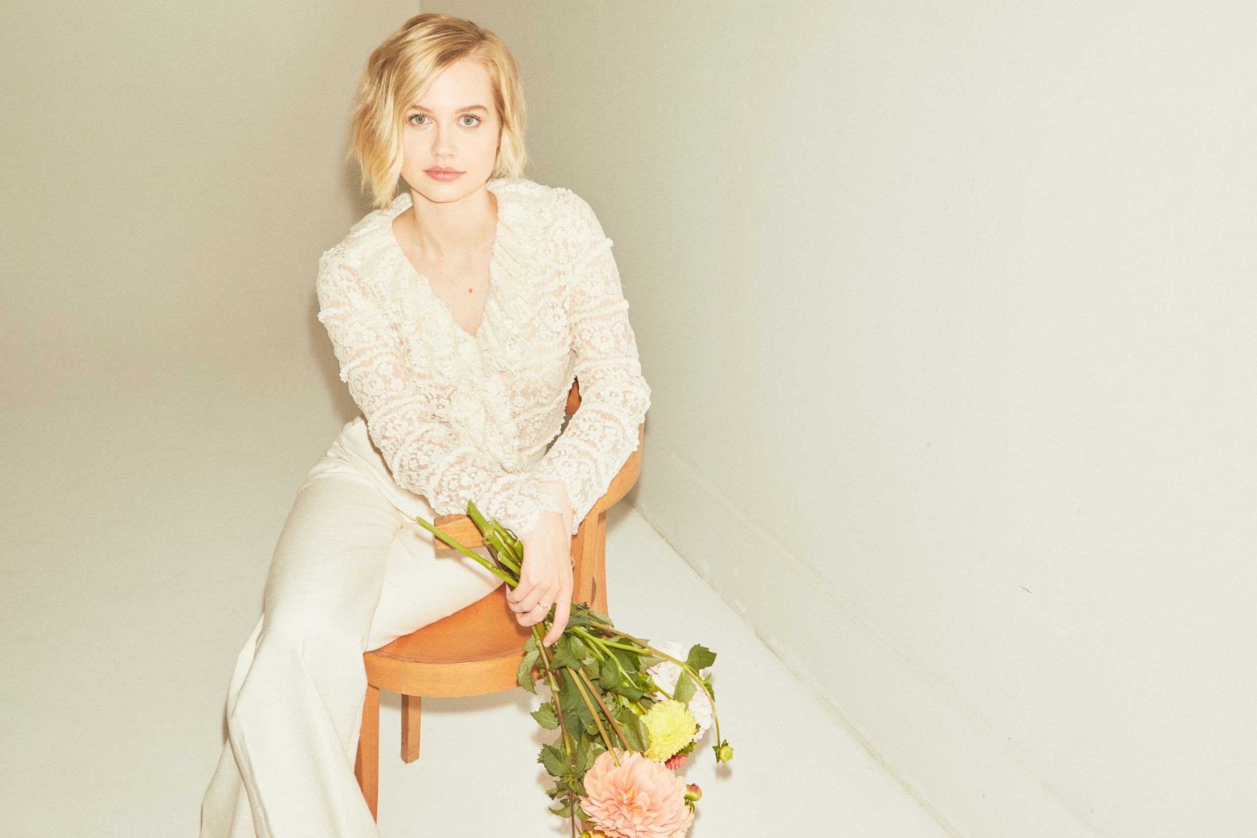 Young woman with short blonde hair poses with a bouquet while sitting on a wooden chair in front of a white wall in a photo studio.