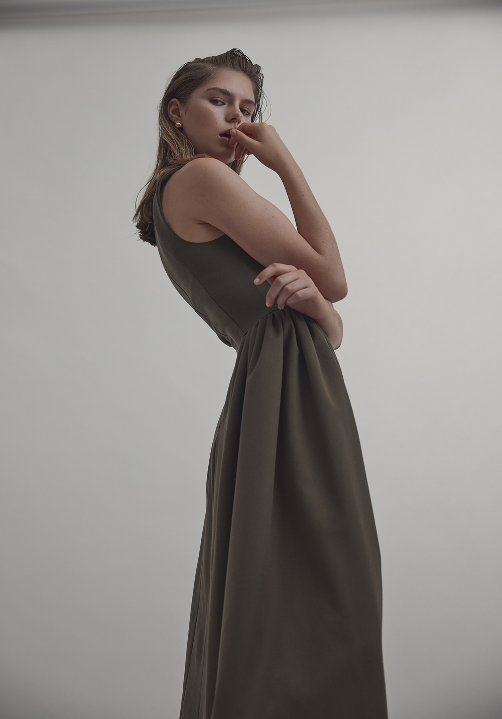 Adult female model in a sleeveless olive-green dress, posing with a hand near her mouth in a studio against a white background.