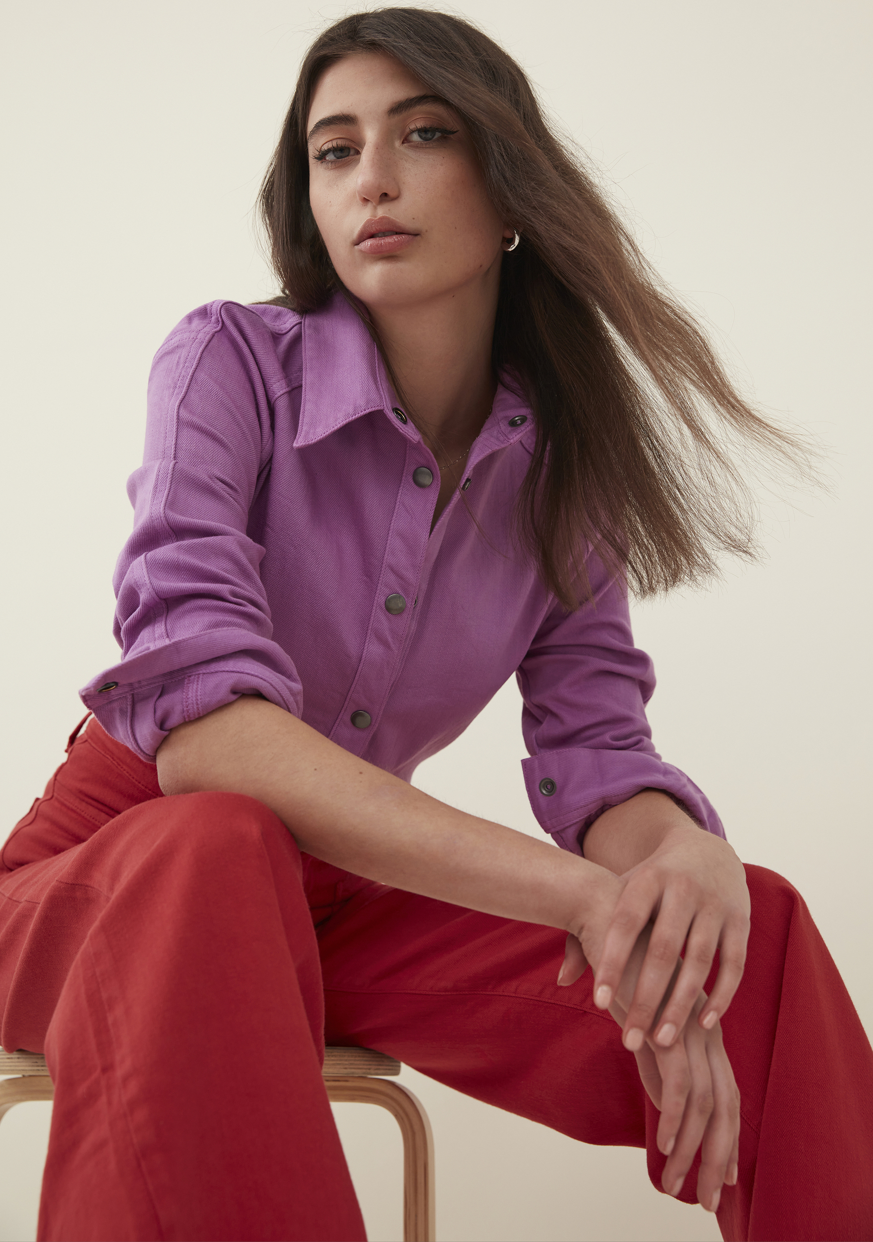 Indoor portrait of a woman in a purple shirt and red pants sitting on a stool looking at the camera with a neutral expression.