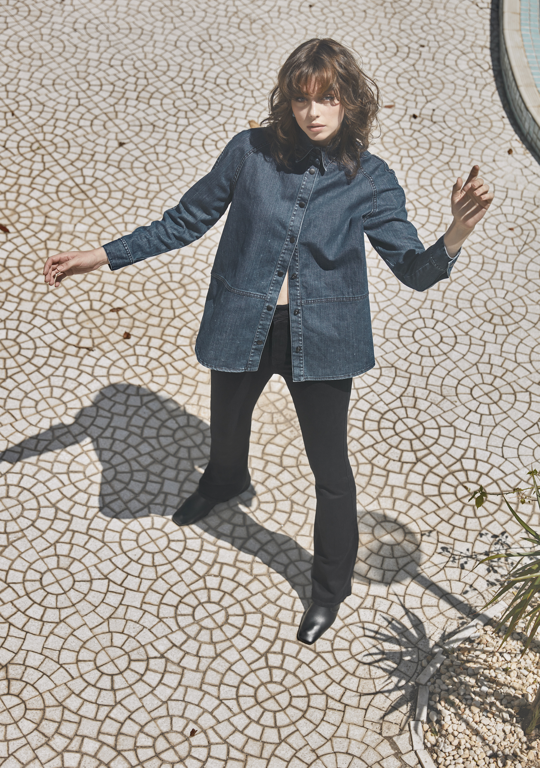 A woman in a denim shirt and black pants stands on a patterned tile surface in an outdoor setting with natural lighting and shadows.