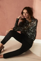 Woman in dark floral print blouse and black trousers sitting on a floor against a textured pink wall styled by Sarah Banger.