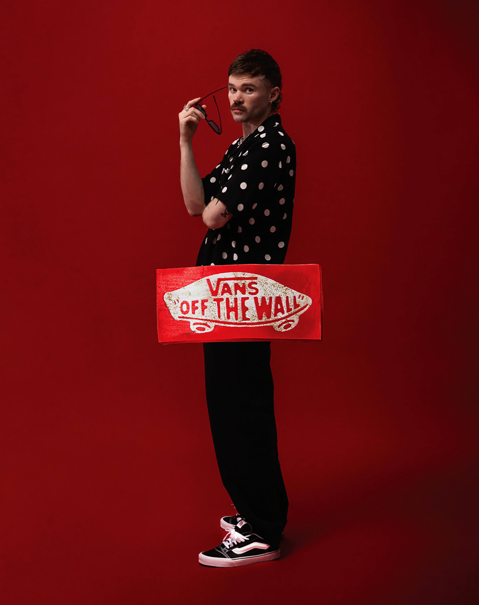 Stylish man poses against red backdrop wearing a polka dot shirt, black pants, and Vans sneakers, holding sunglasses next to a Vans logo board.