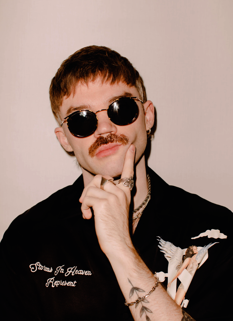Portrait of a man with a mustache wearing round sunglasses and a black shirt with a graphic design, posing indoors.