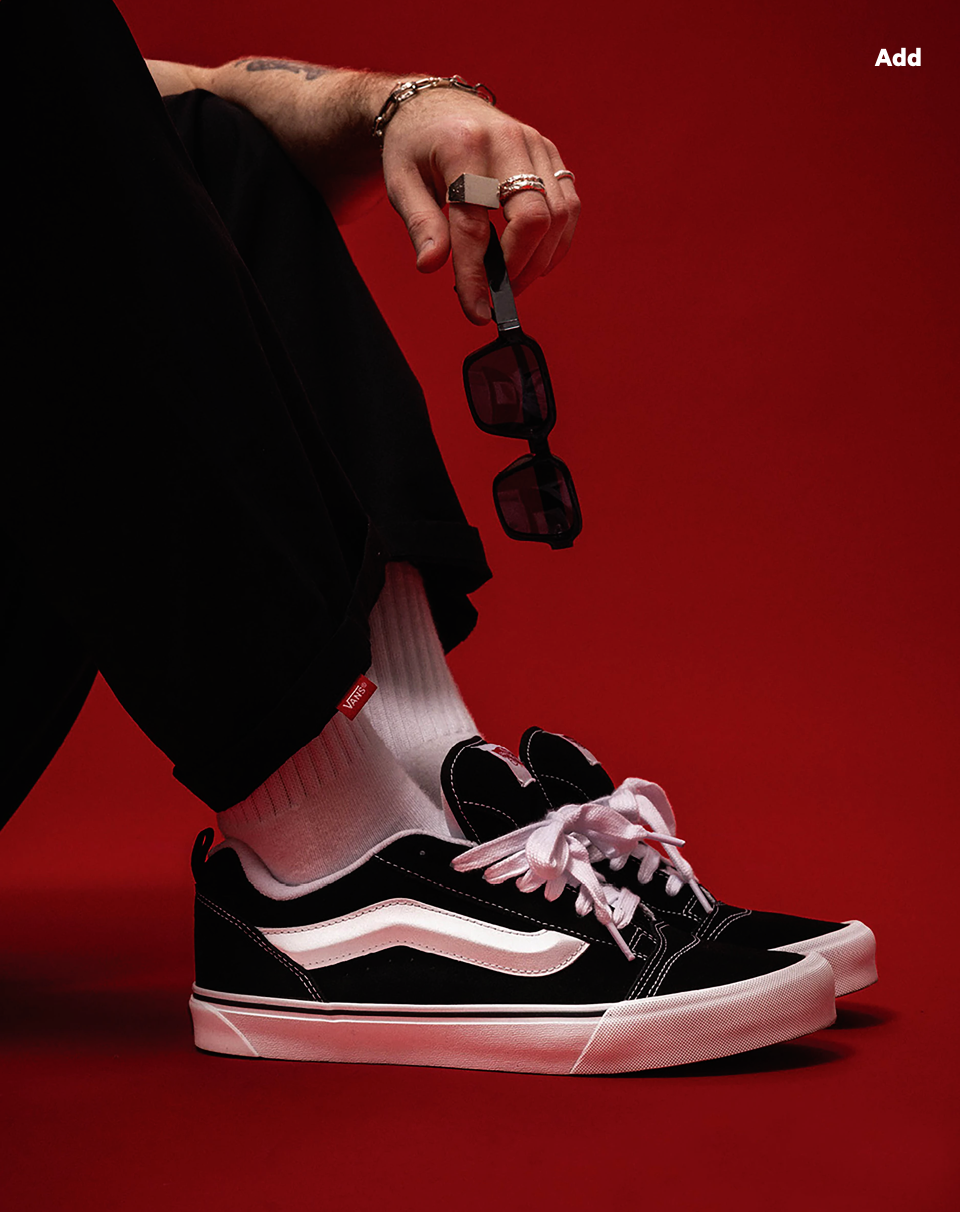 Black and white Vans sneakers on a red surface, worn with white socks and black pants, styled with sunglasses and jewelry.