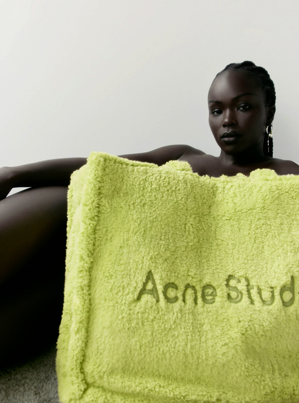 Adult female poses partially covered by a light green towel with 'Acne Studios' logo. Dark skin, dark braided hair. Neutral facial expression.