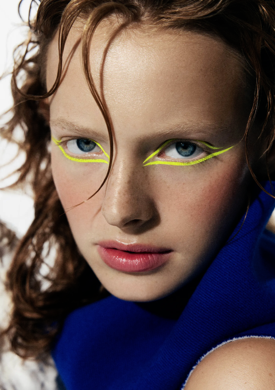 Close-up of a young adult woman with red hair and glowing blue eyes, wearing vibrant yellow graphic eyeliner in a studio setting.