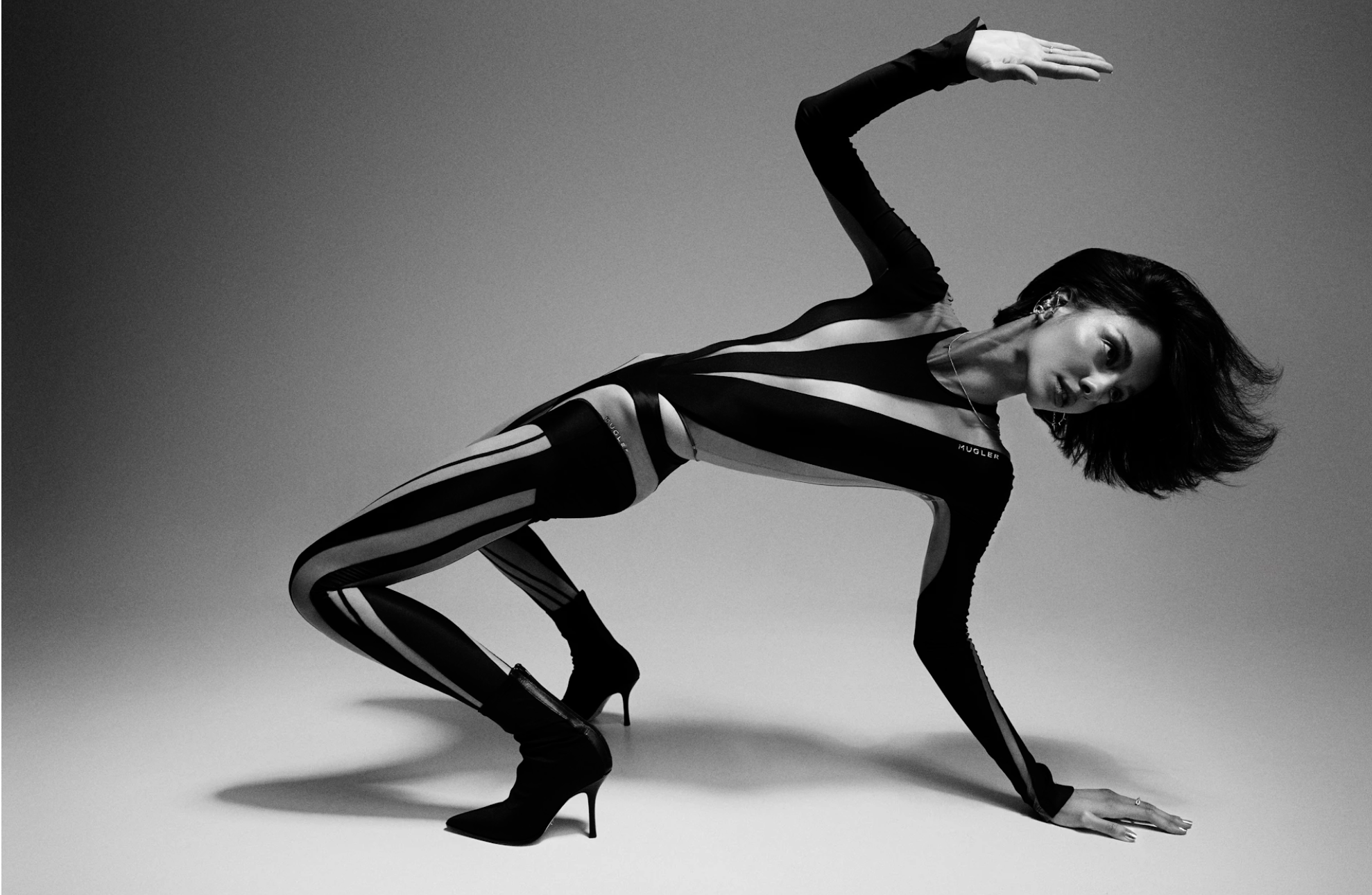 Fashion shot of a female model in a black and white bodysuit striking a dynamic pose on a light gray background with studio lighting.