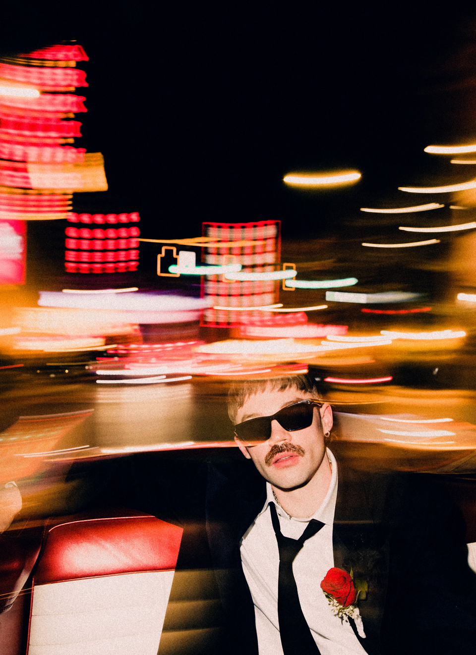 Portrait of a young adult man wearing dark sunglasses, a suit and tie, and a boutonniere while sitting in a car at night.