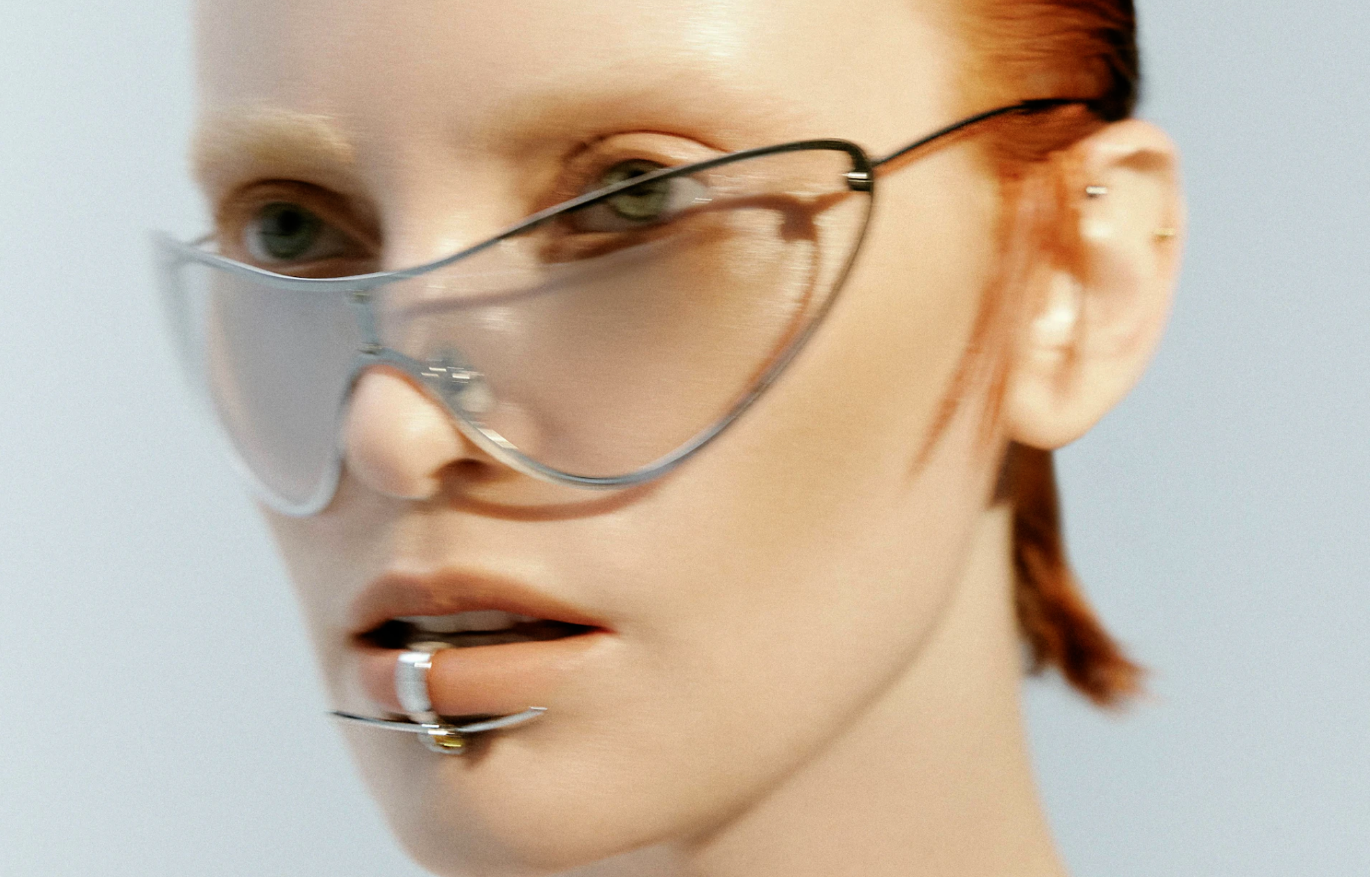 Close-up of a woman with fair skin, red hair, and striking silver sunglasses. She has a lip piercing and a neutral facial expression.