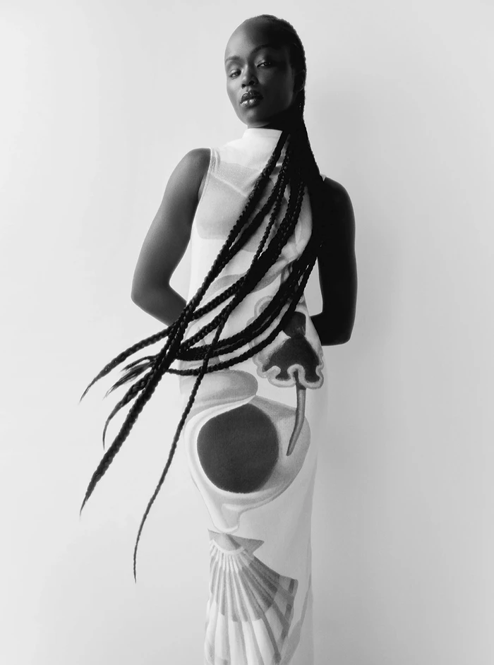 Black and white fashion portrait of a model with long braids wearing a sleeveless patterned dress against a seamless white background.