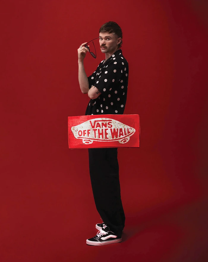 Man holding sunglasses and wearing a black polka dot shirt standing against a solid red background