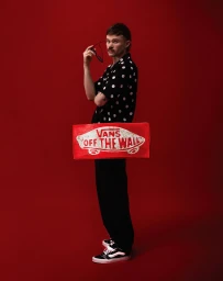 Man holding sunglasses and wearing a black polka dot shirt standing against a solid red background