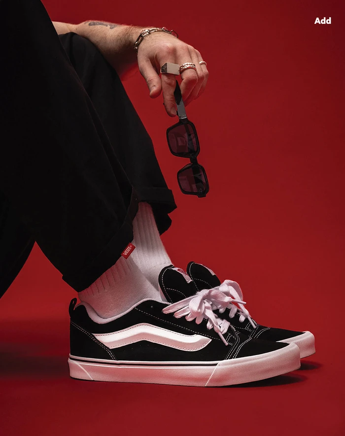 Close-up portrait of a person wearing black sneakers and white socks against a solid red background, holding sunglasses.