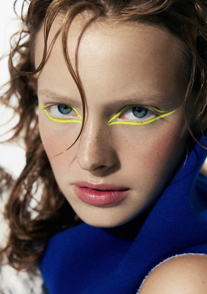 Close-up portrait of a model with blue eyes and striking neon yellow graphic eyeliner, wearing a vibrant royal blue top.