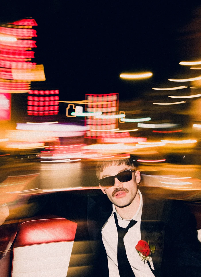 Man wearing sunglasses, black suit, and red rose boutonniere in a car with blurred city lights outside.