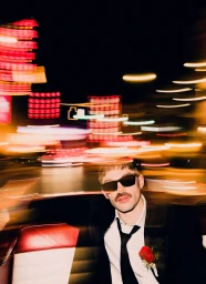 Man wearing sunglasses, black suit, and red rose boutonniere in a car with blurred city lights outside.
