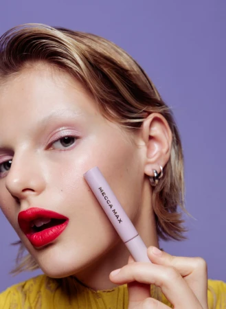 Young adult woman with red lipstick holds a MECCA MAX product tube in a studio shot against a purple background. Beauty concept.