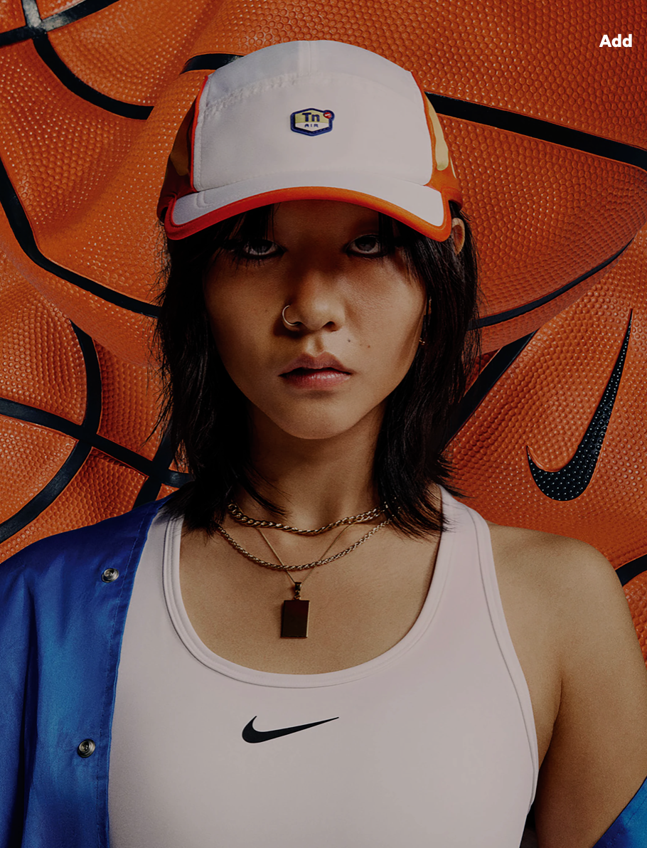 Portrait of a young woman in a white Nike sports bra and cap, with layered gold necklaces, against a background of orange basketballs.