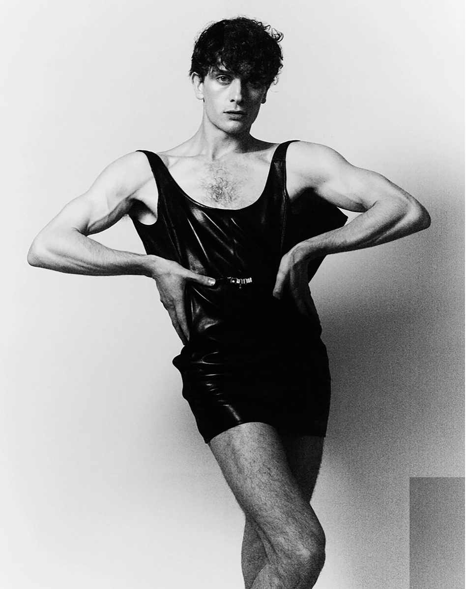 Monochrome fashion portrait of a male model posing in a black, shiny, sleeveless top and shorts in a photo studio setting.