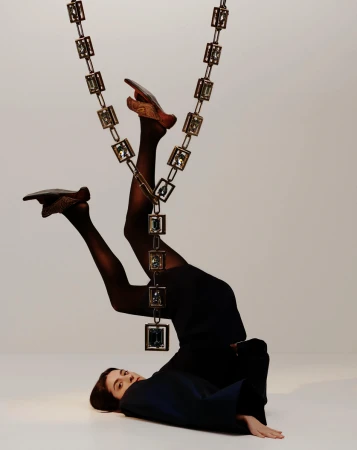 Model lying upside down on a white seamless floor with legs suspended upward by an ornate jeweled chain.