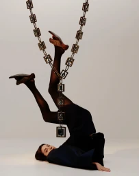 Model lying upside down on a white seamless floor with legs suspended upward by an ornate jeweled chain.