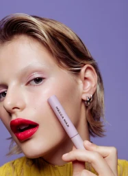Close-up beauty portrait of a woman with red lipstick holding a cosmetic tube near her cheek against a purple background
