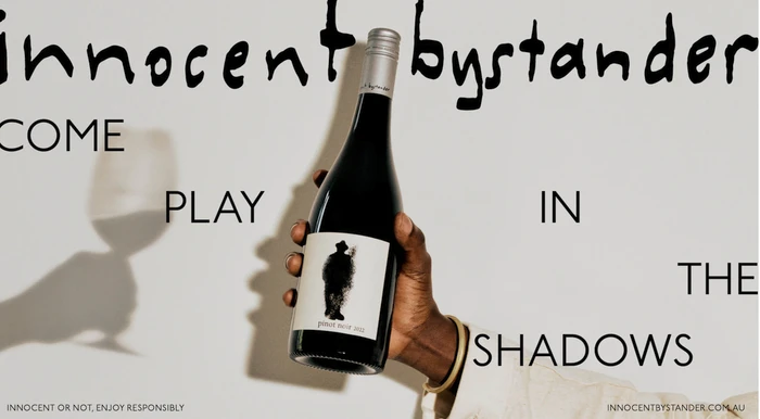Close-up of a person holding a dark wine bottle against a plain background with bold typography and a wine glass shadow