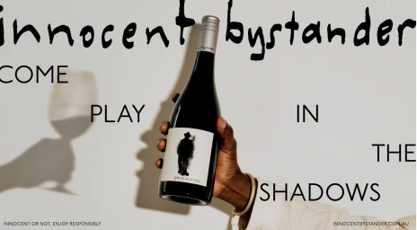 Close-up of a person holding a dark wine bottle against a plain background with bold typography and a wine glass shadow