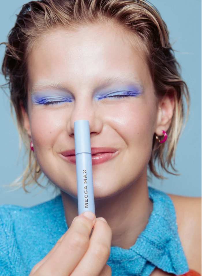Close-up portrait of a smiling woman with blue eyeshadow holding a blue cosmetic tube vertically against her nose