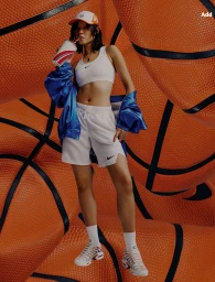 Woman in white sports bra and shorts drinking from a cup, posed against giant textured basketballs.