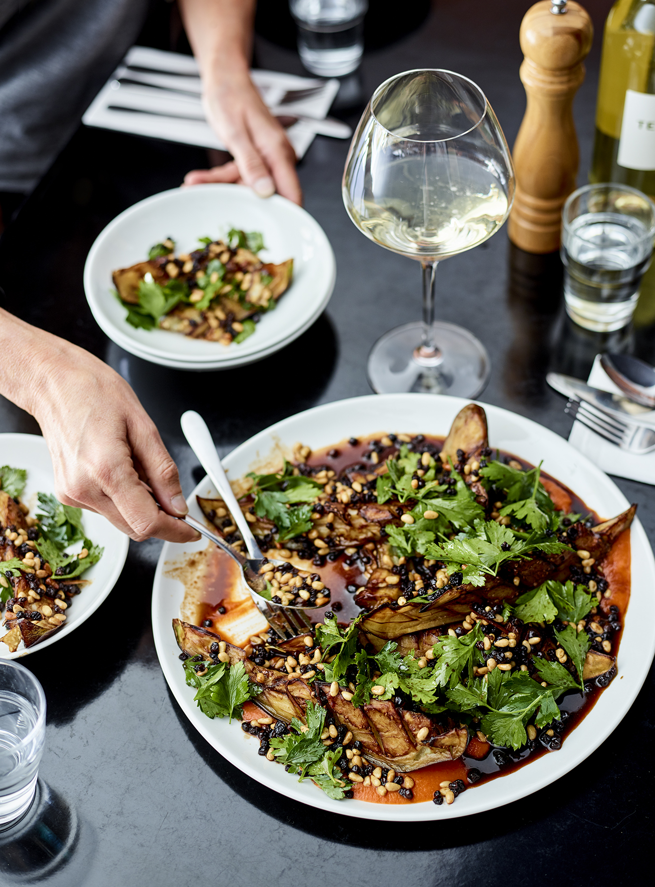 Overhead shot of a table setting with a large salad with greens, potatoes, and sauce, served on white plates alongside a glass of wine.