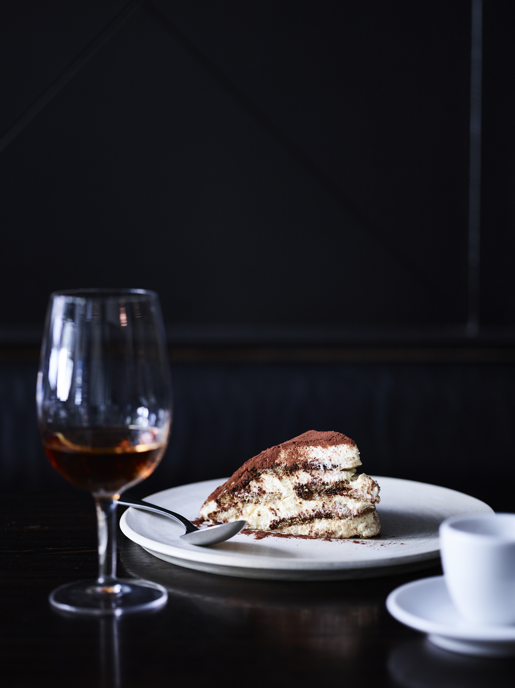 Tiramisu dessert slice on a white plate with a spoon, a glass of amber-colored liquid, and a white coffee cup on a dark table.