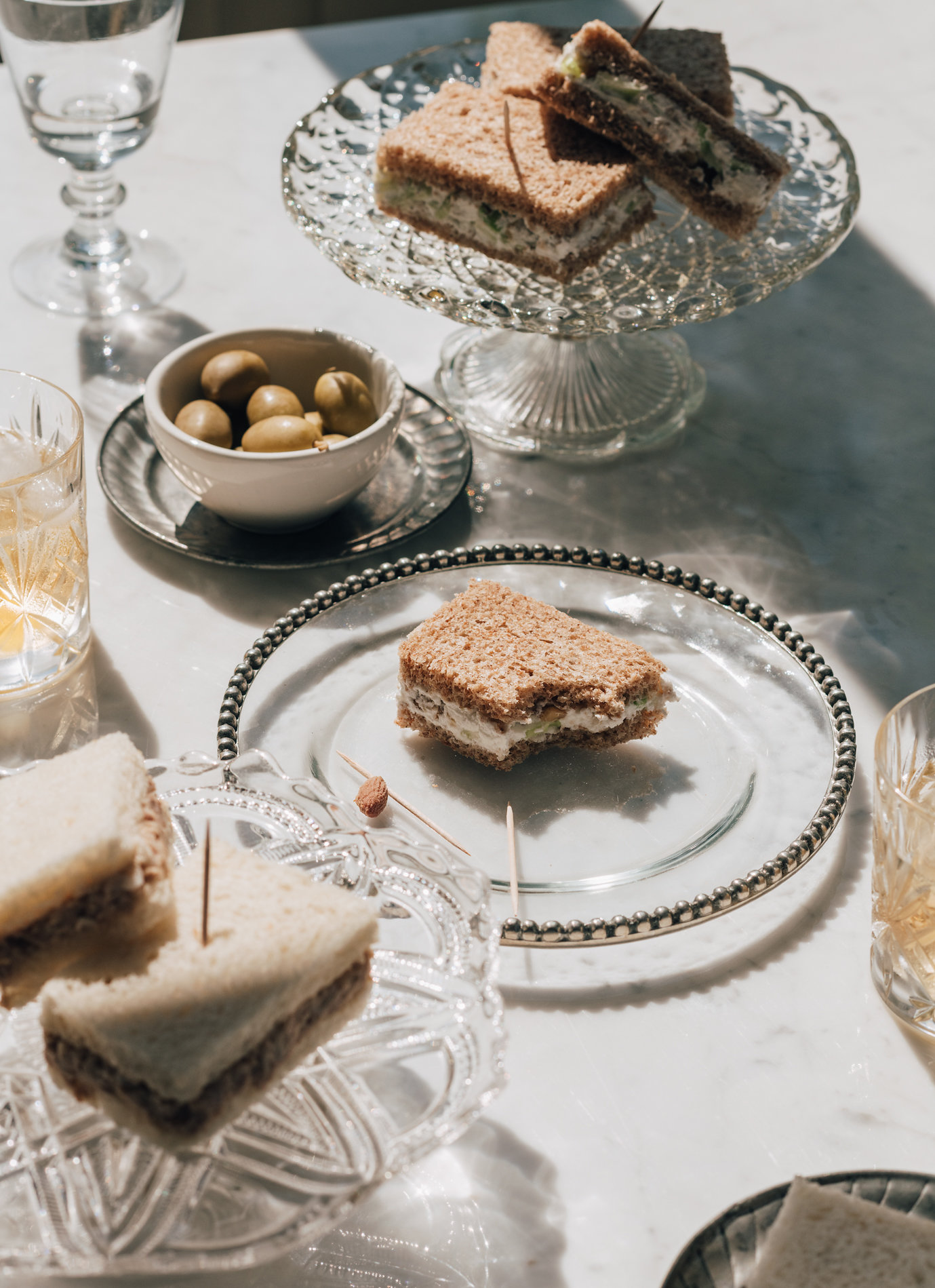 Sandwich squares with toothpicks served on decorative glass plates alongside a bowl of green olives and cut-crystal glassware.