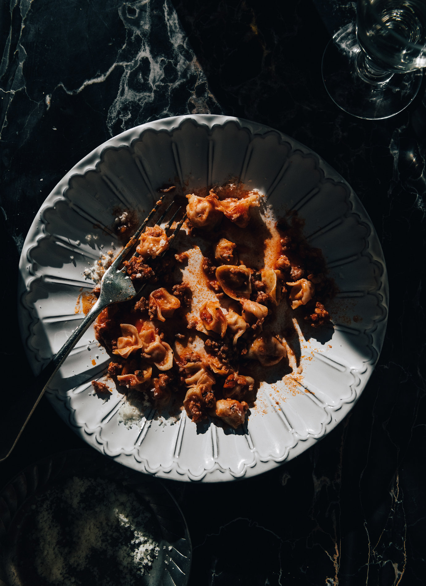 Overhead shot of a white plate with leftover pasta with sauce and a fork on a dark marble table next to a glass of water.