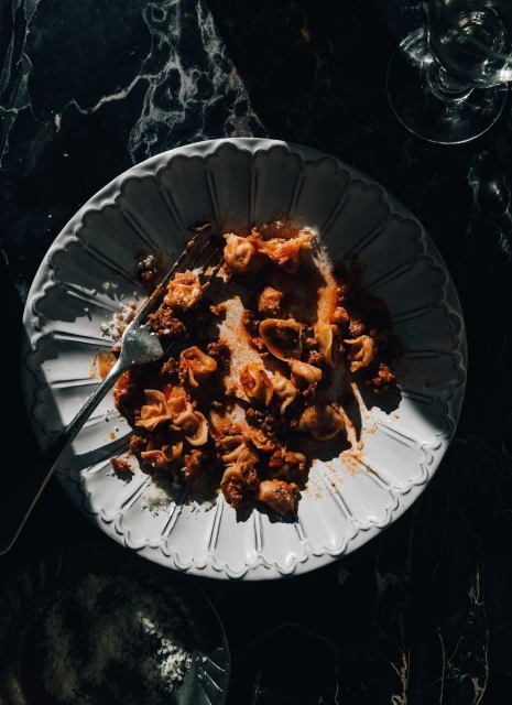 Overhead shot of a white plate with leftover pasta with sauce and a fork on a dark marble table next to a glass of water.